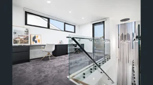 Modern home office with large windows, a white chair at a dark desk, framed artwork, and grey carpet. A sleek glass balustrade with metal handrail overlooks the staircase with hanging pendant lights. Natural light fills the space.