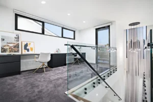 Modern home office with large windows, a white chair at a dark desk, framed artwork, and grey carpet. A sleek glass balustrade with metal handrail overlooks the staircase with hanging pendant lights. Natural light fills the space.