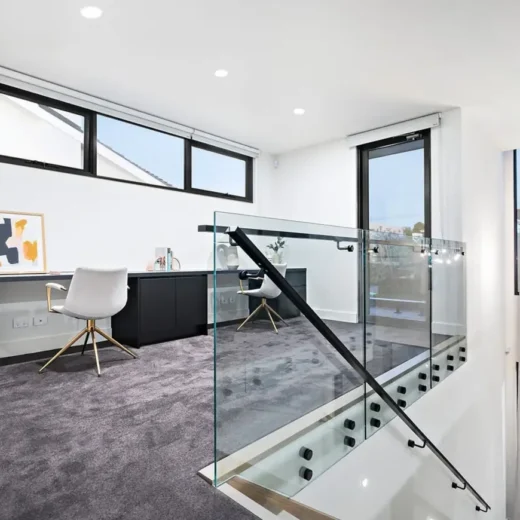 Glass Balustrade Installation across Melbourne Modern home office with large windows, a white chair at a dark desk, framed artwork, and grey carpet. A sleek glass balustrade with metal handrail overlooks the staircase with hanging pendant lights. Natural light fills the space.